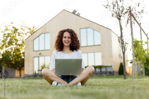 Принти на полотні Full length frontal portrait of a smiling young woman with curly hair with laptop seated on grass in the her yard