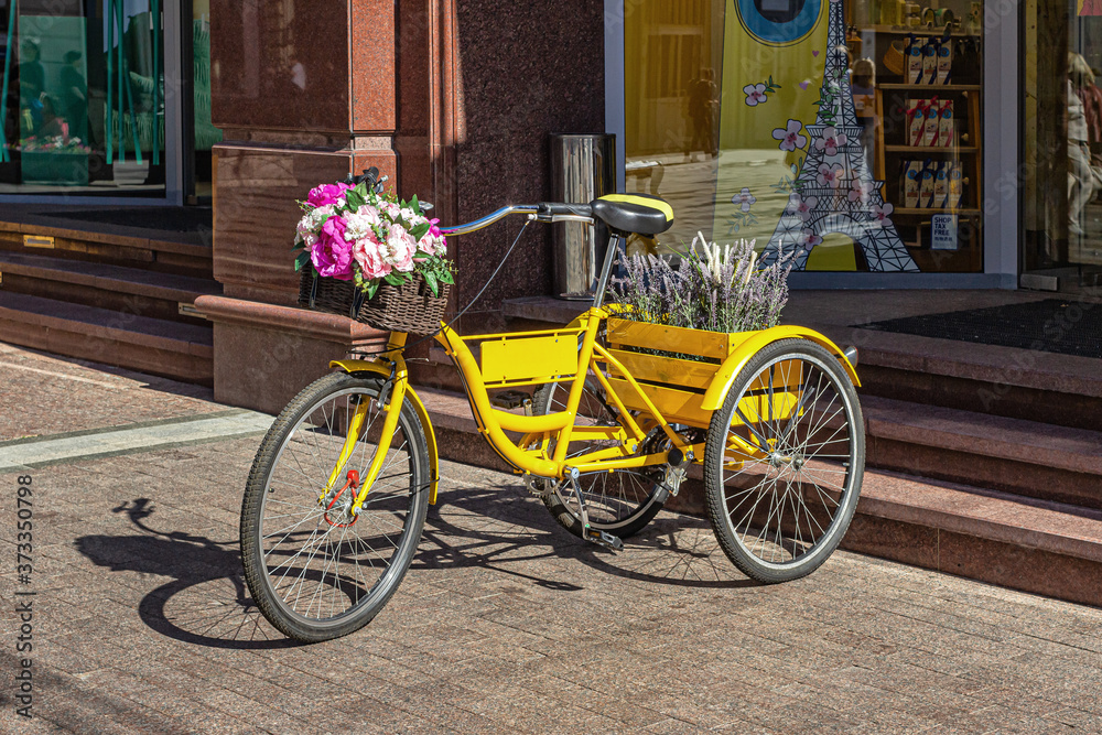 Yellow tricycle with flowers in baskets, standing at the entrance to the store