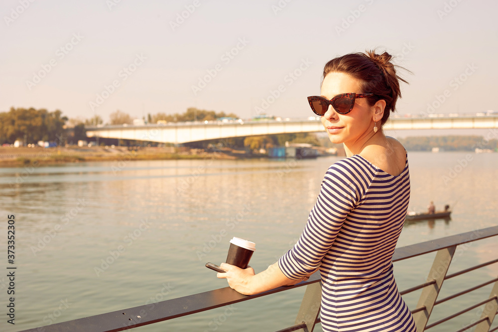 Beautiful woman using a cellphone while drinking coffee near a river.