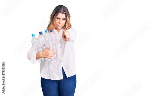 Young caucasian woman holding recycling plastic bottles pointing with finger to the camera and to you, confident gesture looking serious