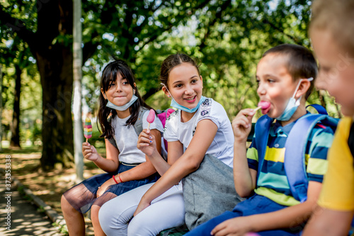 Group of children eating ice cream  outside. They are wearing a protective face mask down. © Ana