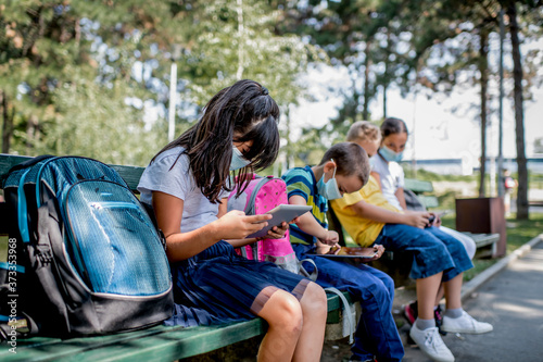 Back to school during quarantine. Children with backpacks wearing protective face mask and using mobile phones and digital tablets. © Ana