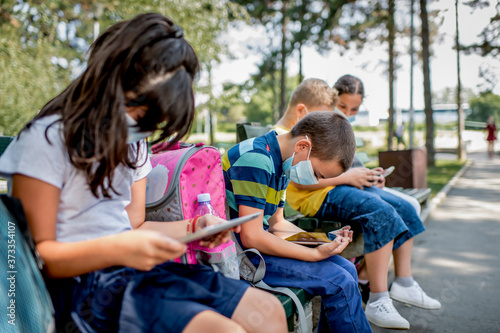 Back to school during quarantine. Children with backpacks wearing protective face mask and using mobile phones and digital tablets.