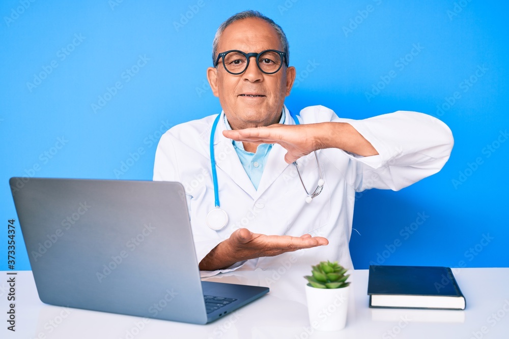 Senior handsome man with gray hair wearing doctor uniform working using computer laptop gesturing with hands showing big and large size sign, measure symbol. smiling looking at the camera. measuring. Senior handsome man with gray hair wearing doctor uniform working using computer laptop gesturing with hands showing big and large size sign, measure symbol. smiling looking at the camera. measuring.