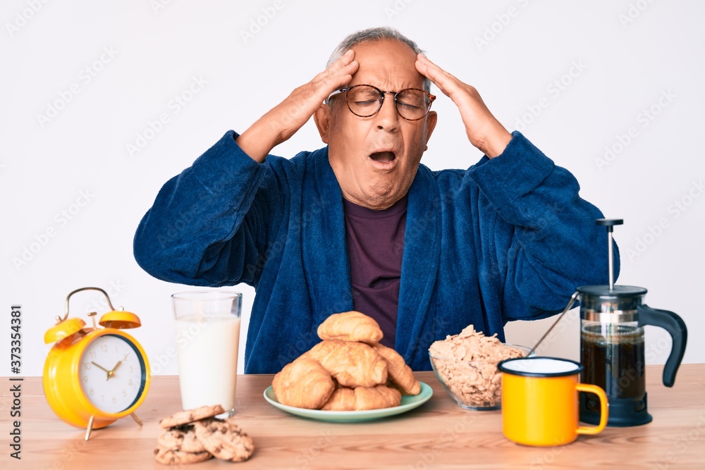 Senior handsome man with gray hair sitting on the table eating breakfast in the morning with hand on head, headache because stress. suffering migraine. Senior handsome man with gray hair sitting on the table eating breakfast in the morning with hand on head, headache because stress. suffering migraine.