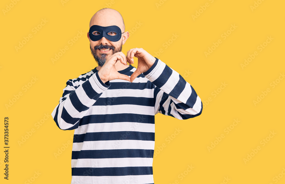 Young handsome man wearing burglar mask smiling in love doing heart symbol shape with hands. romantic concept. Young handsome man wearing burglar mask smiling in love doing heart symbol shape with hands. romantic concept.
