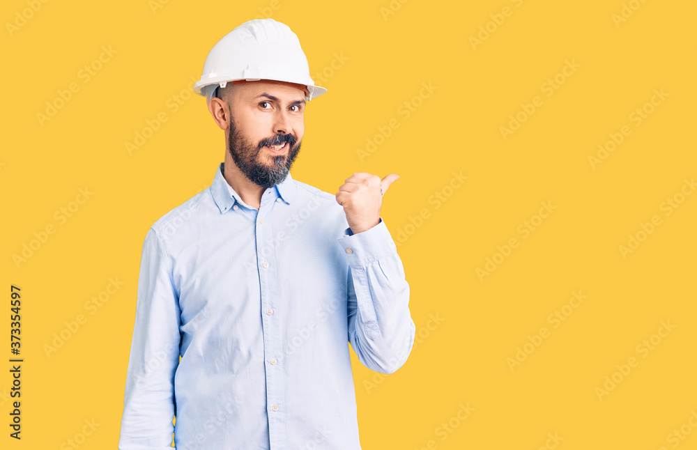 Young handsome man wearing architect hardhat smiling with happy face looking and pointing to the side with thumb up. Young handsome man wearing architect hardhat smiling with happy face looking and pointing to the side with thumb up.