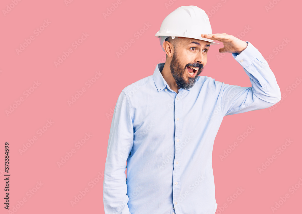 Young handsome man wearing architect hardhat very happy and smiling looking far away with hand over head. searching concept.