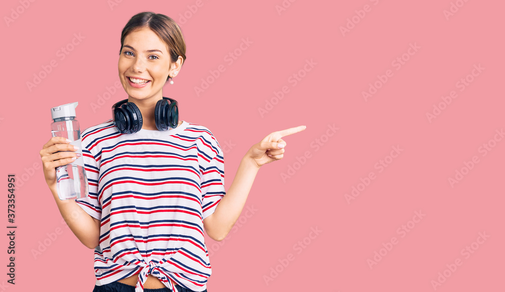 Beautiful caucasian woman with blonde hair wearing sportswear and headphones drinking a bottle of water smiling happy pointing with hand and finger to the side Beautiful caucasian woman with blonde hair wearing sportswear and headphones drinking a bottle of water smiling happy pointing with hand and finger to the side