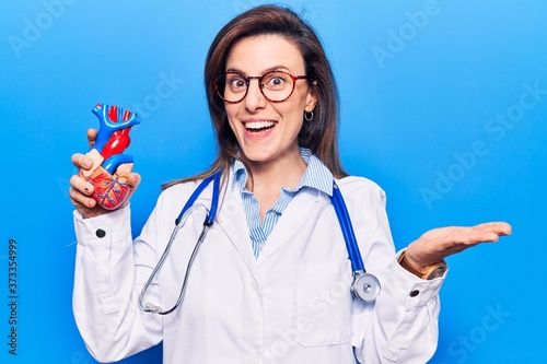 Young beautiful woman wearing doctor stethoscope holding heart celebrating achievement with happy smile and winner expression with raised hand