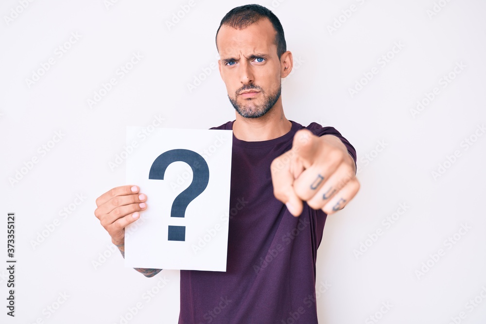 Young handsome man with tattoo holding question mark pointing with finger to the camera and to you, confident gesture looking serious Young handsome man with tattoo holding question mark pointing with finger to the camera and to you, confident gesture looking serious