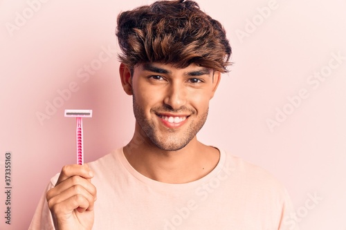 Young hispanic man holding razor looking positive and happy standing and smiling with a confident smile showing teeth