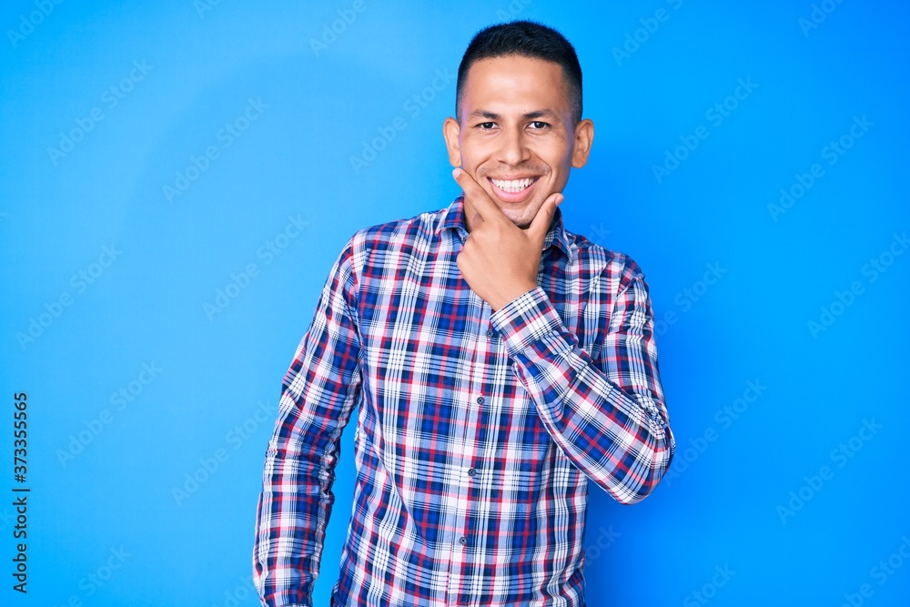 Young handsome latin man wearing casual clothes looking confident at the camera smiling with crossed arms and hand raised on chin. thinking positive.