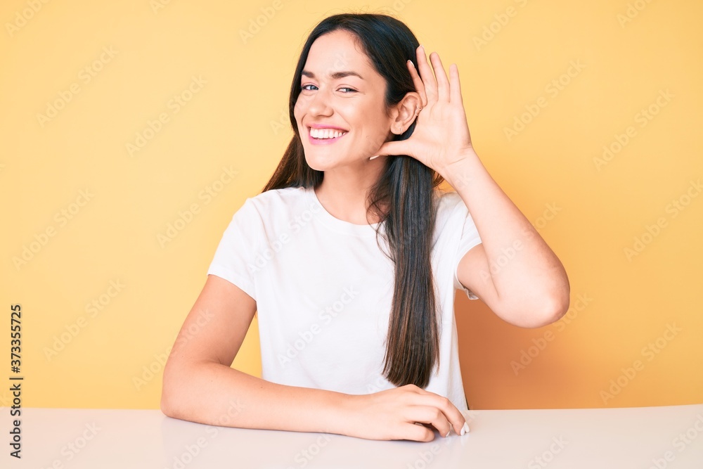Young caucasian woman sitting at the desk wearing casual white tshirt smiling with hand over ear listening and hearing to rumor or gossip. deafness concept. Young caucasian woman sitting at the desk wearing casual white tshirt smiling with hand over ear listening and hearing to rumor or gossip. deafness concept.