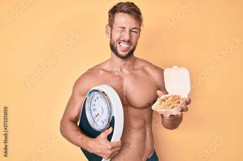 Young caucasian man shirtless holding weighing machine and fried potatoes winking looking at the camera with sexy expression  cheerful and happy face.