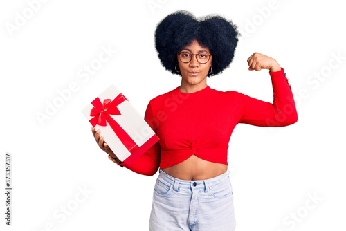 Young african american girl holding gift strong person showing arm muscle, confident and proud of power