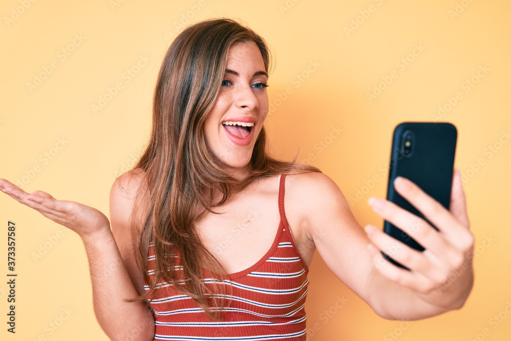 Beautiful young caucasian woman taking a selfie photo with smartphone celebrating achievement with happy smile and winner expression with raised hand Beautiful young caucasian woman taking a selfie photo with smartphone celebrating achievement with happy smile and winner expression with raised hand