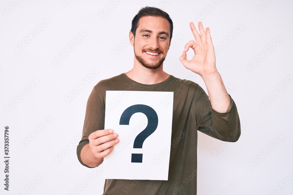 Young handsome man holding question mark doing ok sign with fingers, smiling friendly gesturing excellent symbol Young handsome man holding question mark doing ok sign with fingers, smiling friendly gesturing excellent symbol