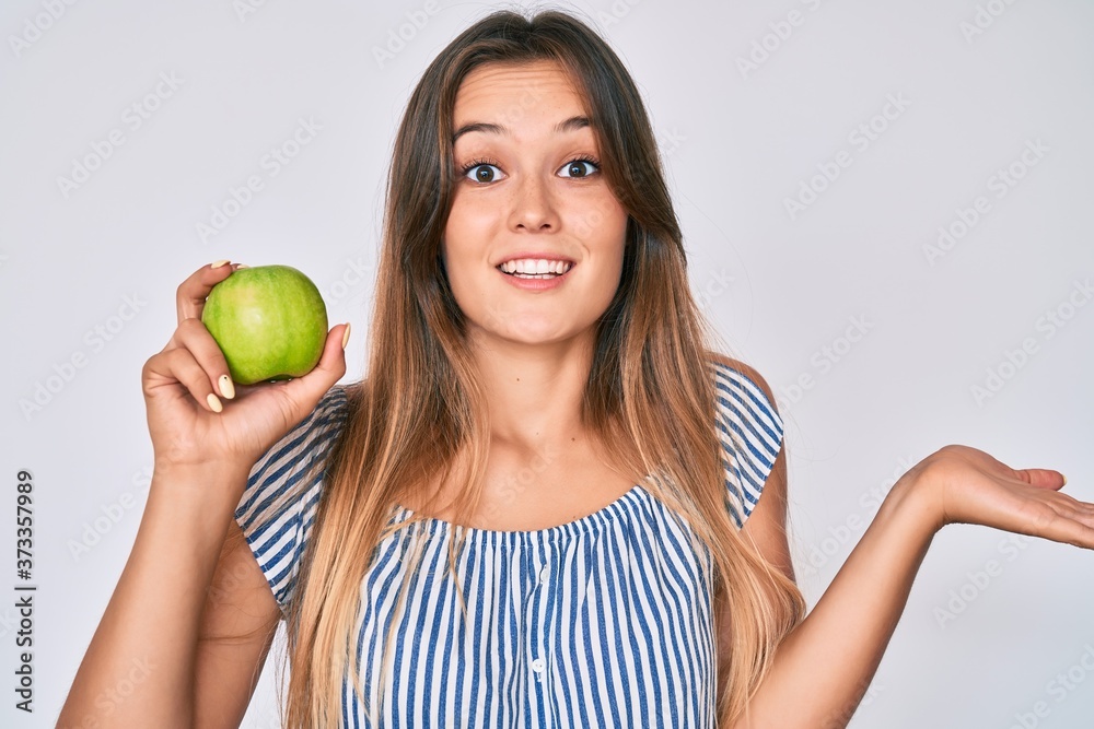 Beautiful caucasian woman holding green apple celebrating achievement with happy smile and winner expression with raised hand Beautiful caucasian woman holding green apple celebrating achievement with happy smile and winner expression with raised hand