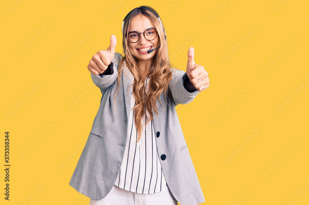 Young beautiful blonde woman wearing call center agent headset approving doing positive gesture with hand, thumbs up smiling and happy for success. winner gesture. Young beautiful blonde woman wearing call center agent headset approving doing positive gesture with hand, thumbs up smiling and happy for success. winner gesture.