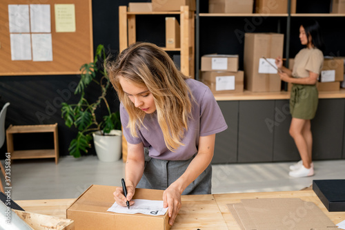 Young serious female manager of online shop writing down address of receiver