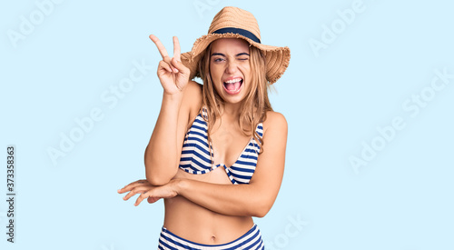Young beautiful blonde woman wearing bikini and hat smiling with happy face winking at the camera doing victory sign with fingers. number two.