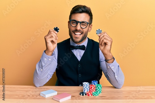 Bilde på lerret Handsome hispanic croupier man sitting on the table holding casino chips winking looking at the camera with sexy expression, cheerful and happy face