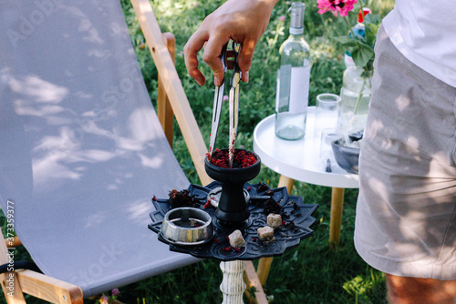 Man's hand distributes red tobacco in a hookah bowl background of summer picnic. Hookah preparation for smoking and fun at summer picnic. Metallic hookah bowl with charcoal inside. 