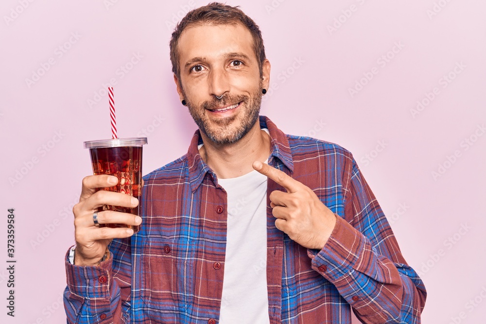 Young handsome man drinking glass of cola beverage smiling happy pointing with hand and finger Young handsome man drinking glass of cola beverage smiling happy pointing with hand and finger