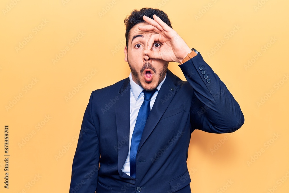 Young hispanic man wearing suit doing ok gesture shocked with surprised face, eye looking through fingers. unbelieving expression. Young hispanic man wearing suit doing ok gesture shocked with surprised face, eye looking through fingers. unbelieving expression.