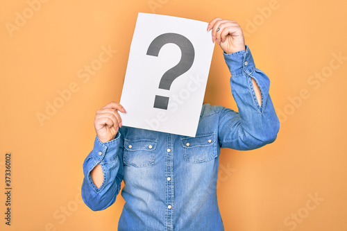 Woman covering face holding paper with question mark symbol. Standing over isolated yellow background