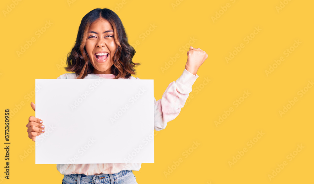 Young beautiful mixed race woman holding cardboard banner with blank space screaming proud, celebrating victory and success very excited with raised arms Young beautiful mixed race woman holding cardboard banner with blank space screaming proud, celebrating victory and success very excited with raised arms