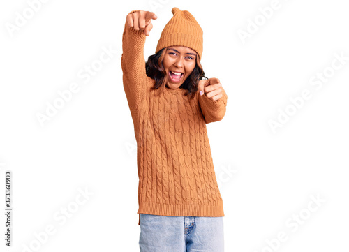 Young beautiful mixed race woman wearing wool sweater and winter hat pointing to you and the camera with fingers, smiling positive and cheerful photo