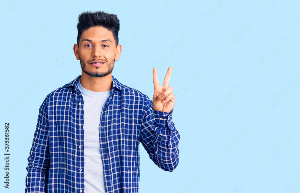 Handsome latin american young man wearing casual shirt smiling with happy face winking at the camera doing victory sign. number two. Handsome latin american young man wearing casual shirt smiling with happy face winking at the camera doing victory sign. number two.