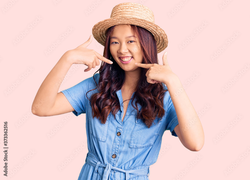 Young beautiful chinese girl wearing summer hat smiling cheerful showing and pointing with fingers teeth and mouth. dental health concept. Young beautiful chinese girl wearing summer hat smiling cheerful showing and pointing with fingers teeth and mouth. dental health concept.