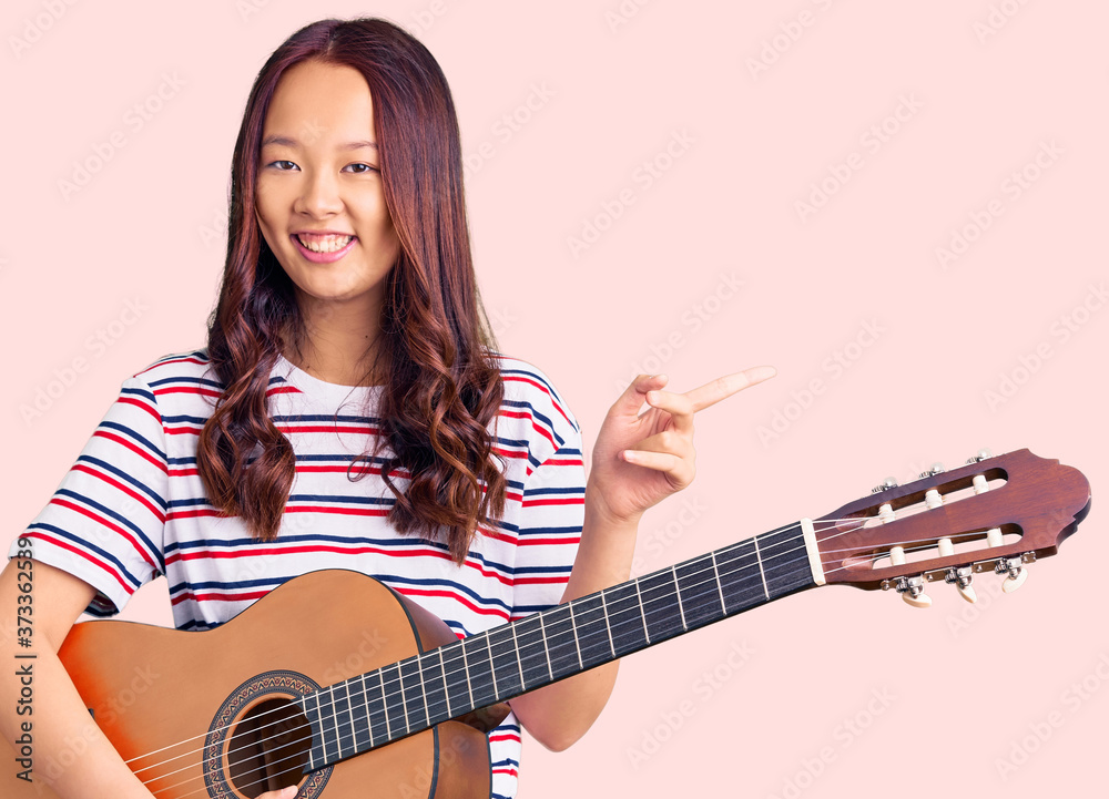 Young beautiful chinese girl playing classical guitar smiling happy pointing with hand and finger to the side