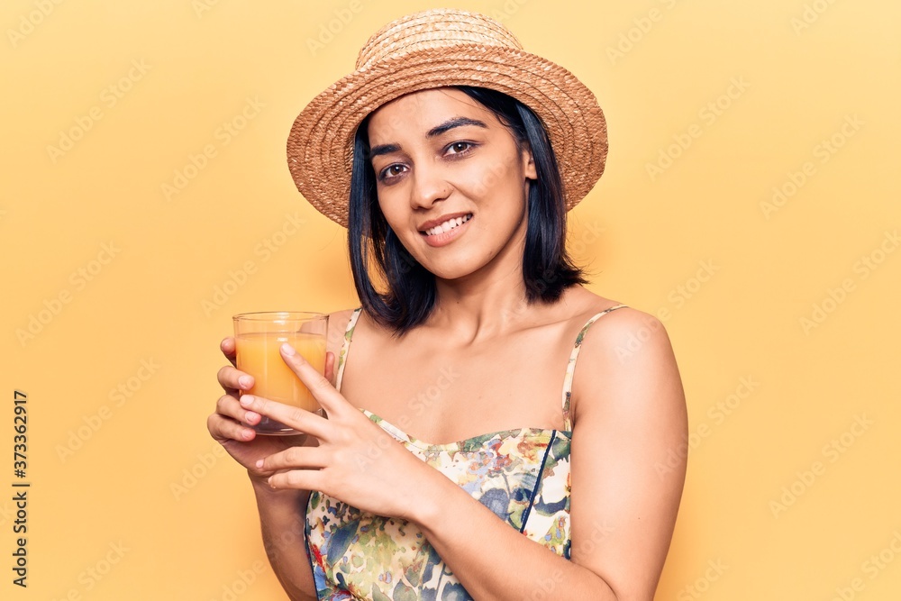 Young beautiful latin woman wearing summer hat drinking glass of orange juice looking positive and happy standing and smiling with a confident smile showing teeth