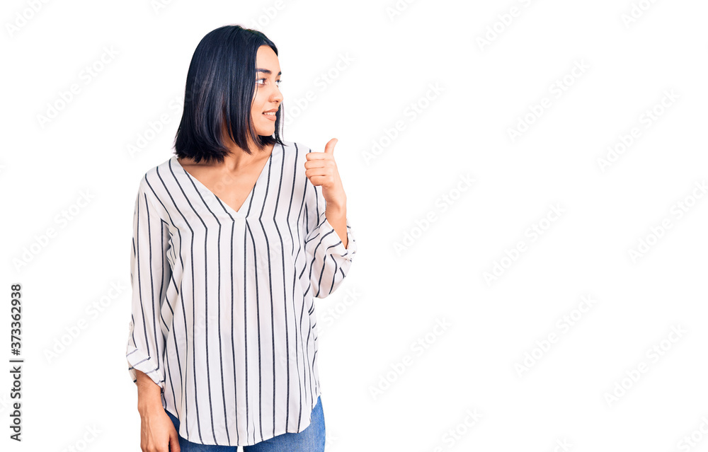 Young beautiful latin woman wearing casual clothes smiling with happy face looking and pointing to the side with thumb up.