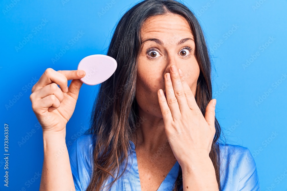 Young beautiful brunette woman applying makeup using sponge over blue background covering mouth with hand, shocked and afraid for mistake. Surprised expression Young beautiful brunette woman applying makeup using sponge over blue background covering mouth with hand, shocked and afraid for mistake. Surprised expression