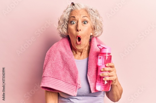 Senior grey-haired woman wearing sportswear and towel drinking bottle of water scared and amazed with open mouth for surprise, disbelief face © Krakenimages.com