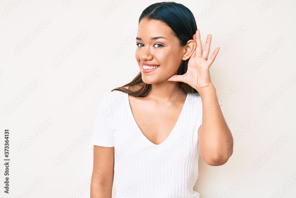 Young beautiful latin girl wearing casual white tshirt smiling with hand over ear listening and hearing to rumor or gossip. deafness concept. Young beautiful latin girl wearing casual white tshirt smiling with hand over ear listening and hearing to rumor or gossip. deafness concept.