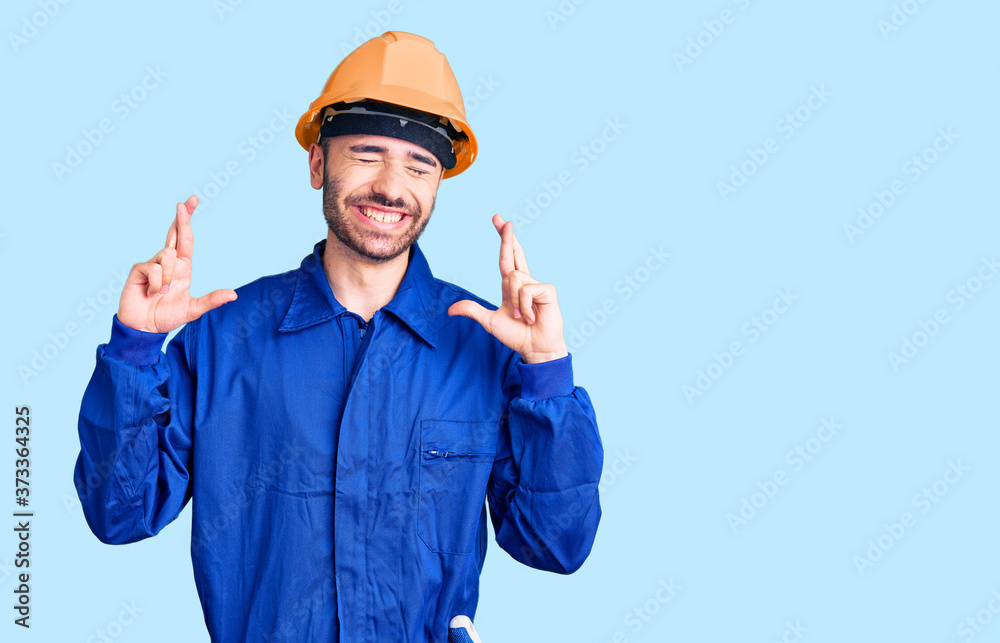 Young hispanic man wearing worker uniform gesturing finger crossed smiling with hope and eyes closed. luck and superstitious concept. Young hispanic man wearing worker uniform gesturing finger crossed smiling with hope and eyes closed. luck and superstitious concept.