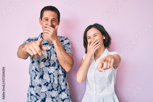 Beautiful couple wearing casual clothes laughing at you, pointing finger to the camera with hand over mouth, shame expression