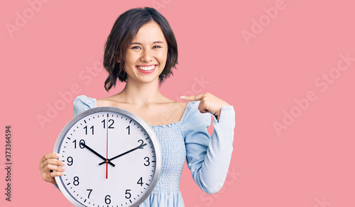 Young beautiful girl holding big clock pointing finger to one self smiling happy and proud