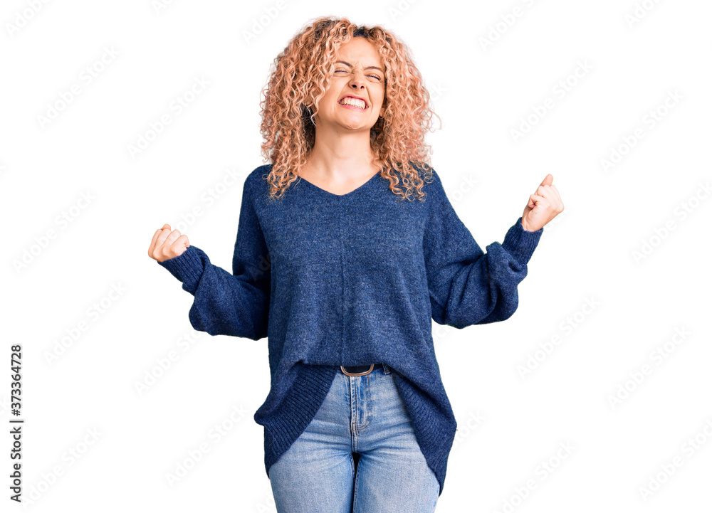 Young blonde woman with curly hair wearing casual winter sweater very happy and excited doing winner gesture with arms raised, smiling and screaming for success. celebration concept. Young blonde woman with curly hair wearing casual winter sweater very happy and excited doing winner gesture with arms raised, smiling and screaming for success. celebration concept.