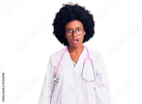 Young african american woman wearing doctor coat and stethoscope in shock face, looking skeptical and sarcastic, surprised with open mouth