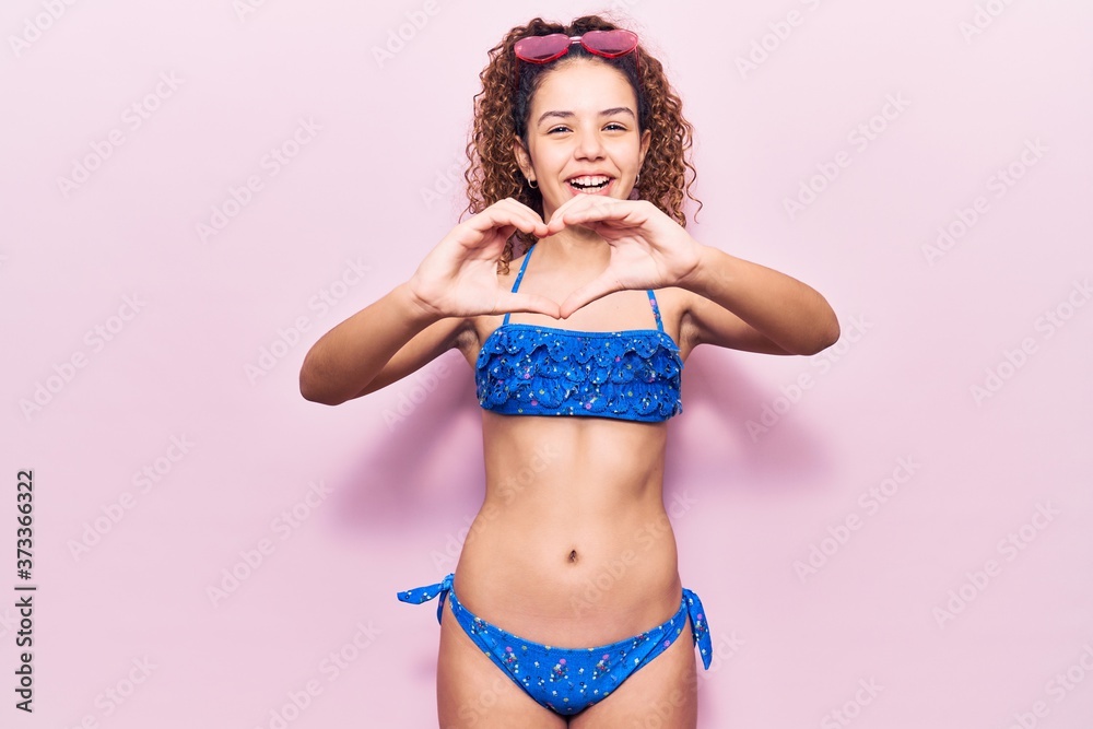Fototapeta premium Beautiful kid girl with curly hair wearing bikini and sunglasses smiling in love doing heart symbol shape with hands. romantic concept.