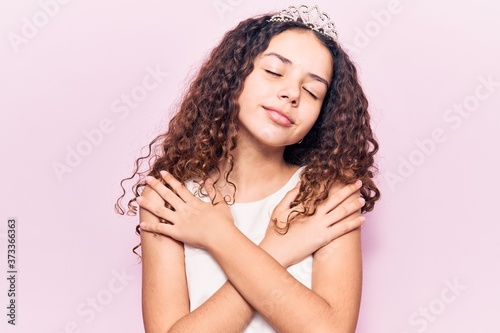 Beautiful kid girl with curly hair wearing princess tiara hugging oneself happy and positive, smiling confident. self love and self care