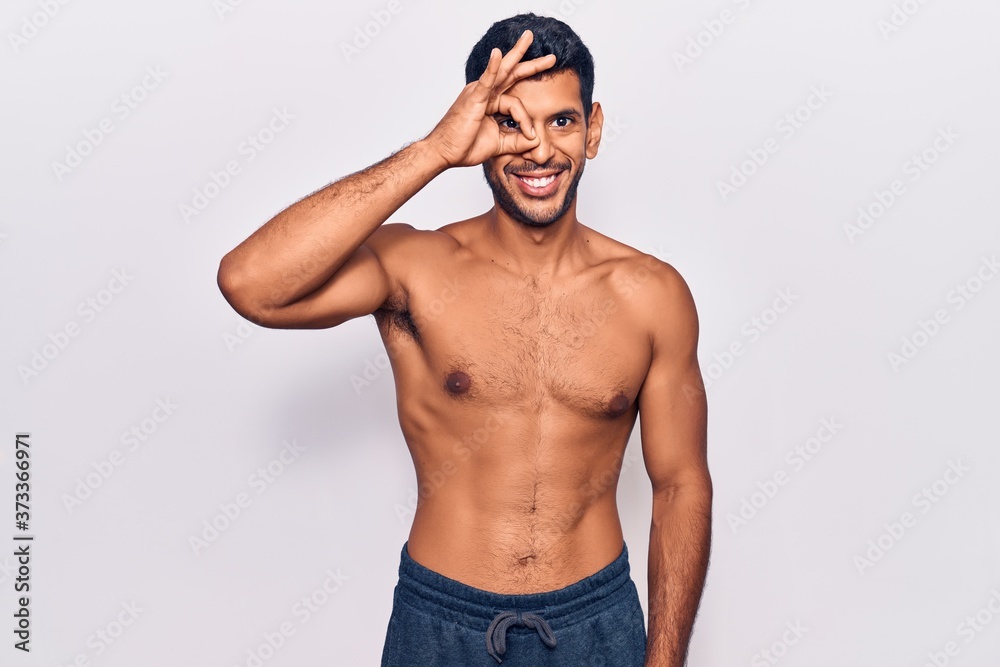 Young latin man standing shirtless doing ok gesture with hand smiling, eye looking through fingers with happy face.