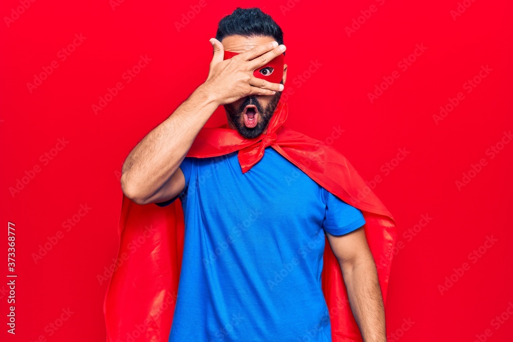 Young hispanic man wearing super hero costume peeking in shock covering face and eyes with hand, looking through fingers afraid Young hispanic man wearing super hero costume peeking in shock covering face and eyes with hand, looking through fingers afraid
